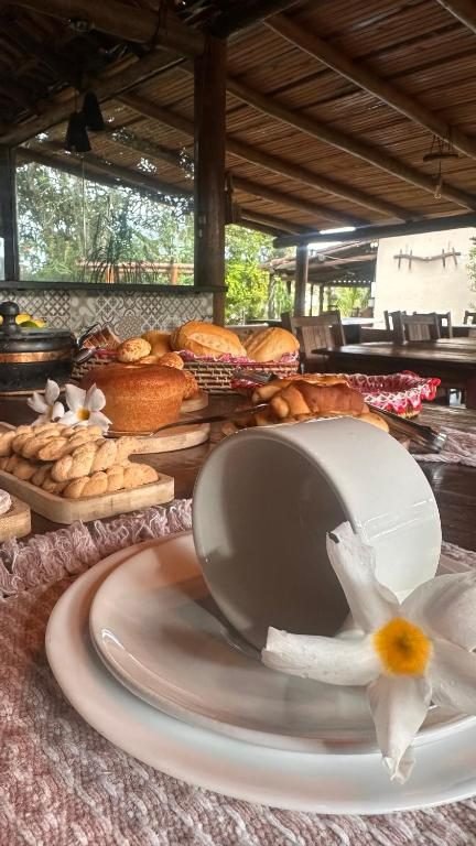 Xícara branca e flor sobre a mesa posta, com pães de queijo e quitandas ao fundo